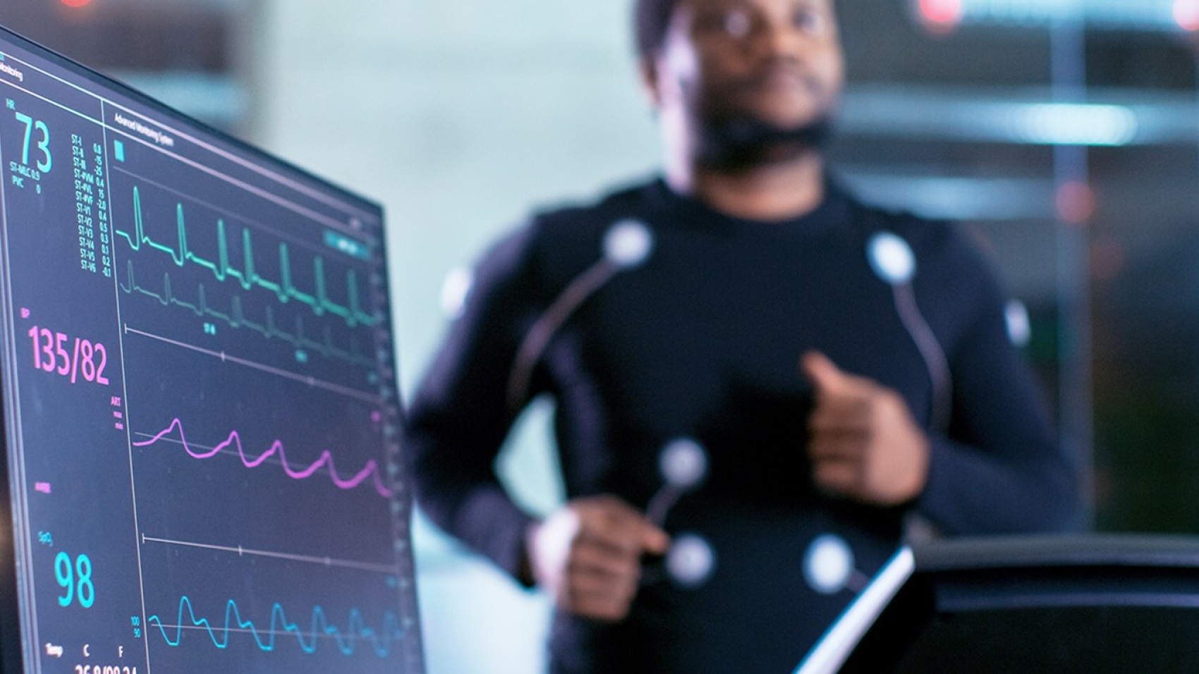 Close-up Shot of a Monitor With EKG Data. Male Athlete Runs on a Treadmill with Electrodes Attached to His Body while Sport Scientist Holds Tablet and Supervises EKG Status in the Background.