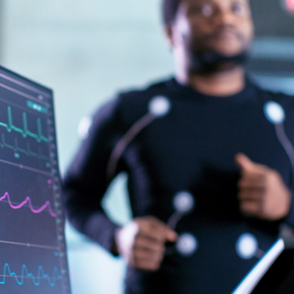 Close-up Shot of a Monitor With EKG Data. Male Athlete Runs on a Treadmill with Electrodes Attached to His Body while Sport Scientist Holds Tablet and Supervises EKG Status in the Background.